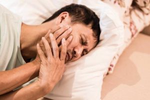 Image of man holding face as he grimaces in pain while laying on a bed.